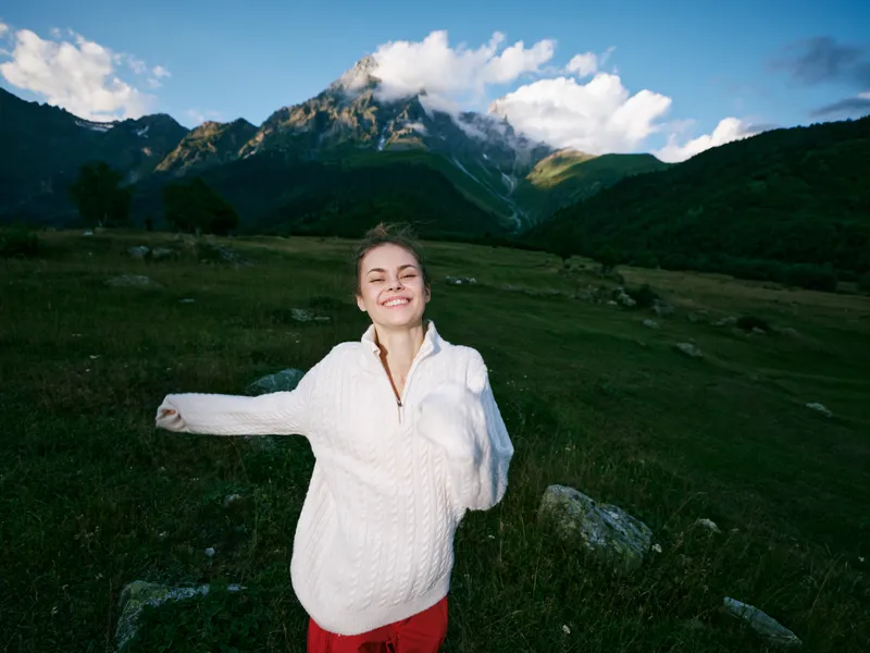 Smiling woman in a white sweater stands in a green meadow with distant mountains, open sky and rolling hills, capturing freedom, nature, and outdoor exploration in bright daylight.