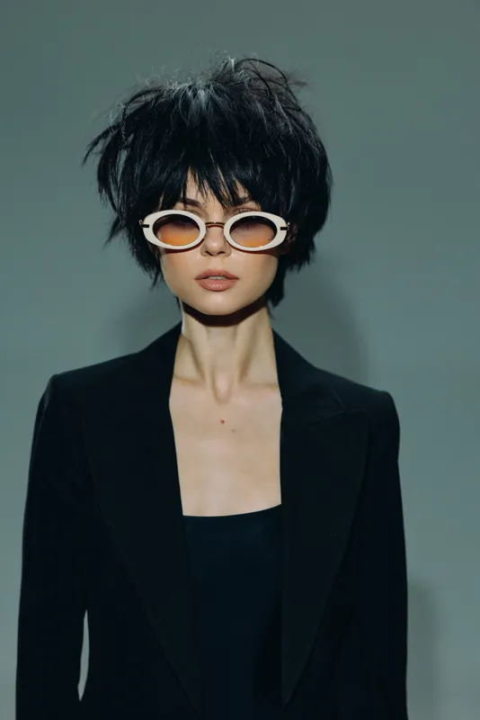 Stylish woman with short black hair wearing white sunglasses and black blazer against gray background