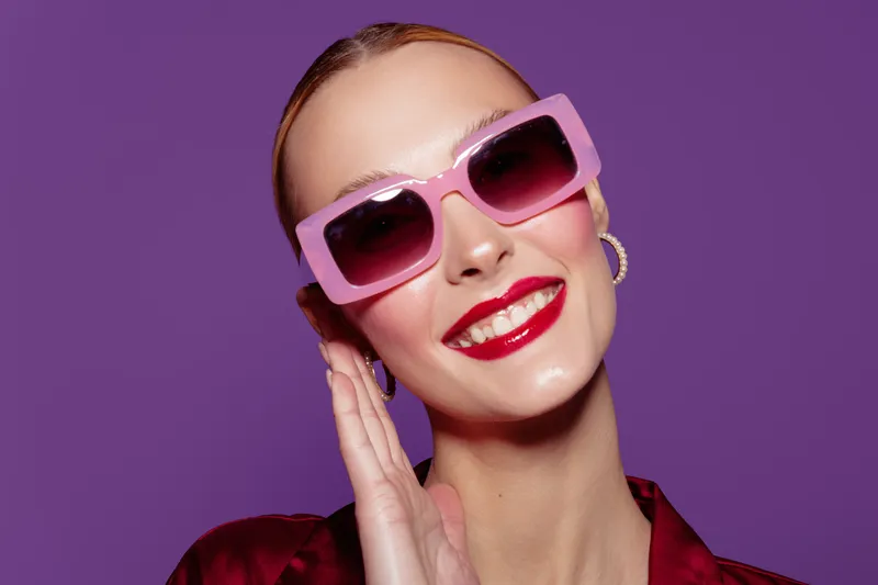 Smiling woman with pink sunglasses and red lipstick poses against purple background. Fashion portrait with bright makeup and stylish accessories. Concept of beauty and confidence.