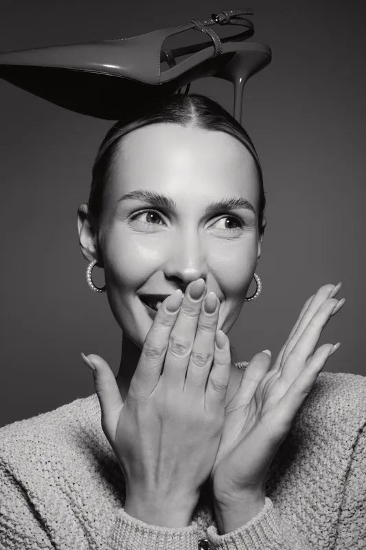 Young caucasian woman with fair skin smiles and covers mouth with hands. She wears hoop earrings and a knitted sweater with a high heel shoe balanced on her head. Creative and playful concept.