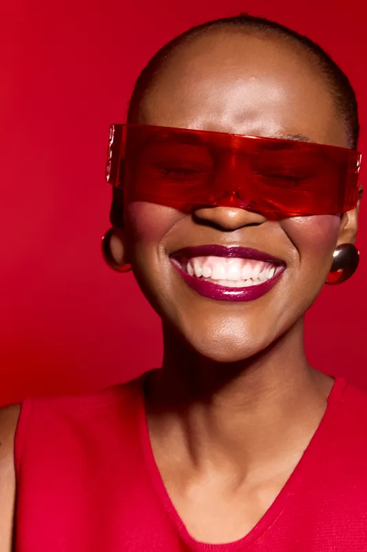 Close-up of a smiling African woman with dark skin wearing red futuristic sunglasses and a red top against a vibrant red background. Concept of fashion, joy, and confidence.