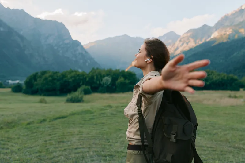A young woman with a backpack stretches her arms in a wide valley while towering mountains rise behind her, conveying a sense of freedom, adventure, and connection with nature.