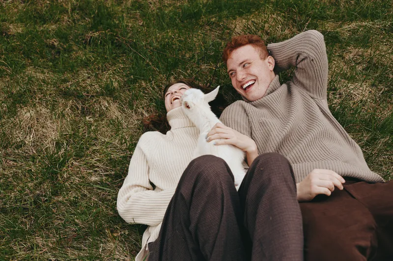 Young couple lying on grass with a baby goat between them laughing and enjoying a relaxed moment in cozy sweaters during a casual outdoor lifestyle scene.