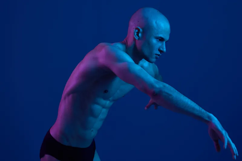 Athletic man posed against a blue background, showcasing muscular physique and dynamic movement. The image utilizes vibrant light to create an energetic atmosphere.