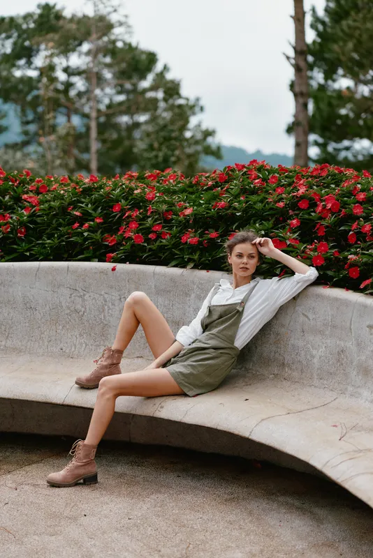 Stylish young model in green overalls sitting on a curved bench, surrounded by vibrant red flowers, exuding confidence and casual elegance