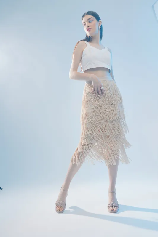 Young woman in a white crop top and beige fringed skirt poses confidently in a bright studio. Concept for woman beauty fashion with elegant and modern style.