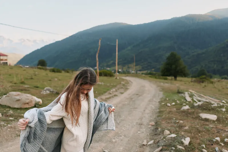 Young woman with long hair wraps a gray shawl around her shoulders while walking on a dirt road in a mountainous rural area during a calm day.