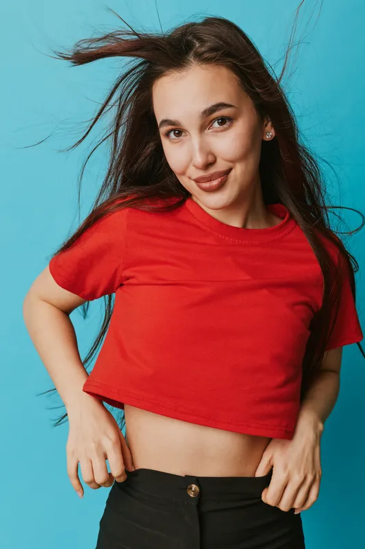 Young woman with long hair wearing red crop top and black pants posing against blue background. Smiling female model with natural makeup and casual style in studio portrait.