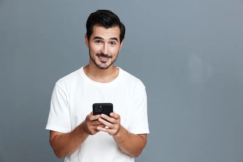 Happy smiling young man wearing a white t-shirt, holding a mobile phone with a friendly expression, posing against a solid grey background. Lifestyle concept.