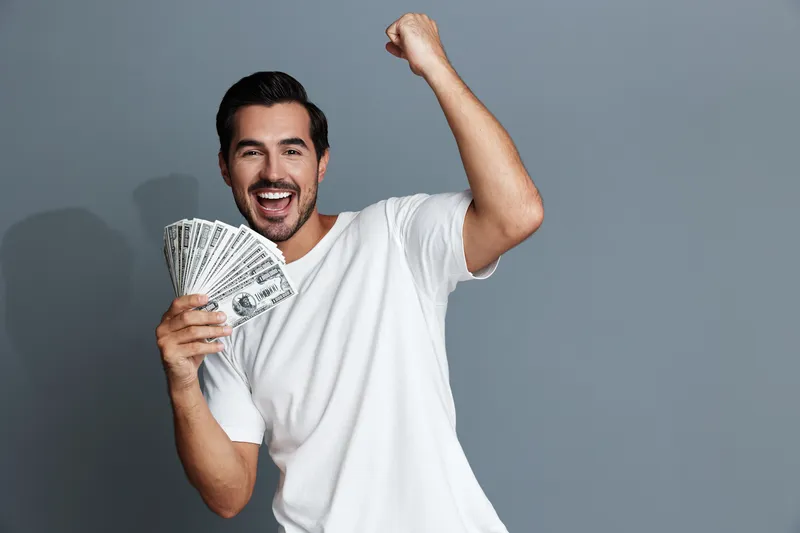 Happy smiling man celebrating success with money in hand, wearing a casual white t-shirt, isolated on solid gray background. Positive emotions and lifestyle concept.