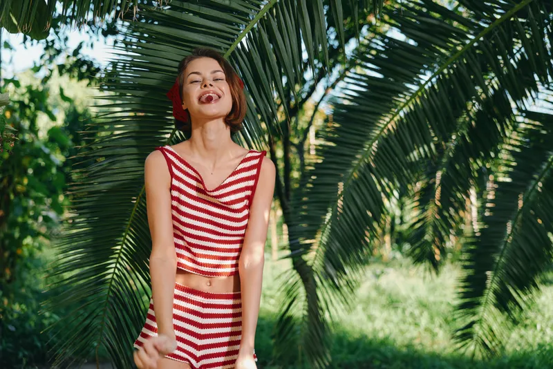 A cheerful woman in a red and white striped bikini stands beneath tropical palms, smiling brightly in a sunny outdoor scene, radiating playful summer energy and carefree mood.