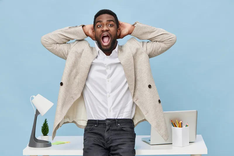 Joyful surprised man with beard, wearing beige blazer and white shirt, standing in bright modern office with laptop, desk lamp, plant, and stationery. People lifestyle concept, expressing shock or exc