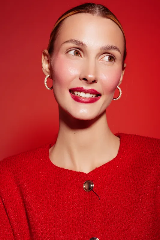Smiling woman with red lipstick and hoop earrings wears red sweater against red background. Close-up portrait with natural skin glow and confident expression. Concept of beauty and fashion.