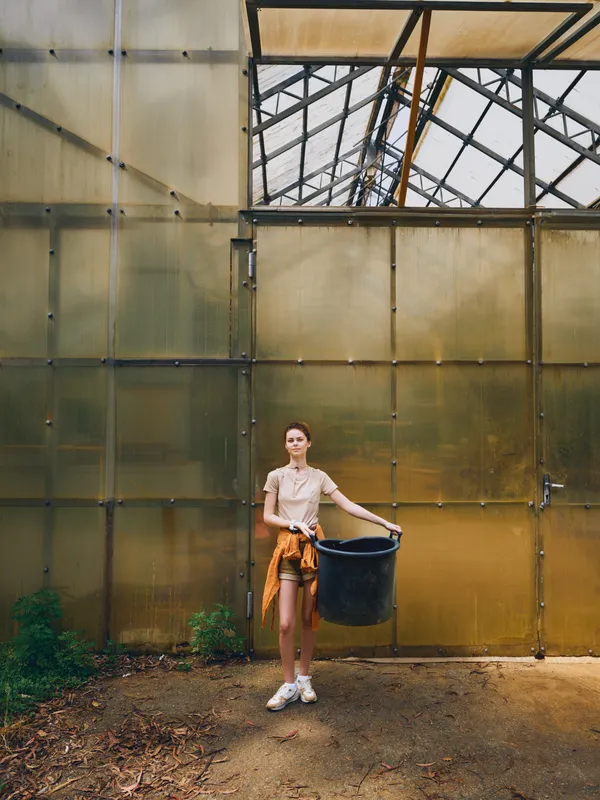 Woman in greenhouse holding a black bucket, gardening and standing near plants in casual clothes, young gardener doing potting and plant care inside a rustic glasshouse with sunlight.