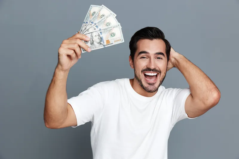 Happy smiling man in white t-shirt holding cash in hand, looking at the camera, isolated on plain solid gray background. Concept of success and wealth.