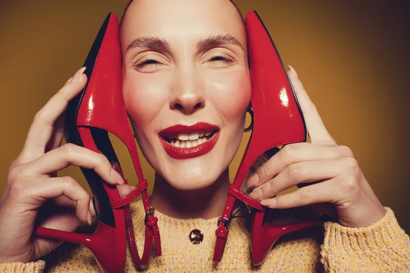 Smiling woman with red lipstick holds red high heel shoes near face, wearing beige sweater. Close-up portrait with warm lighting and joyful expression. Concept of fashion and happiness.