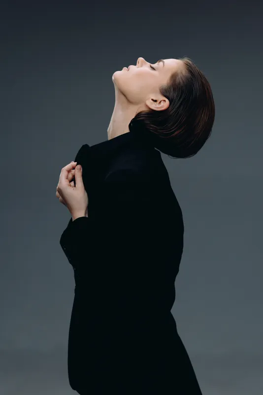 Elegant woman in black coat posing with closed eyes and raised head against dark background.
