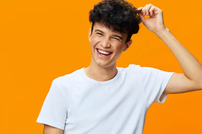 cheerful young man in white t-shirt laughing joyfully against bright orange background with curly hair and casual pose, radiating happiness and warmth