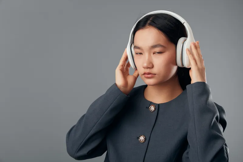 Young woman wearing headphones and closing eyes while listening to music with concentrated expression on gray background. Female headshot with technology and sound concept.