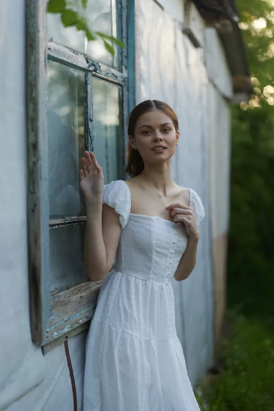 Young woman in a white dress standing by a rustic window, exuding a serene and dreamy emotion in a natural green background, perfect for spring themes