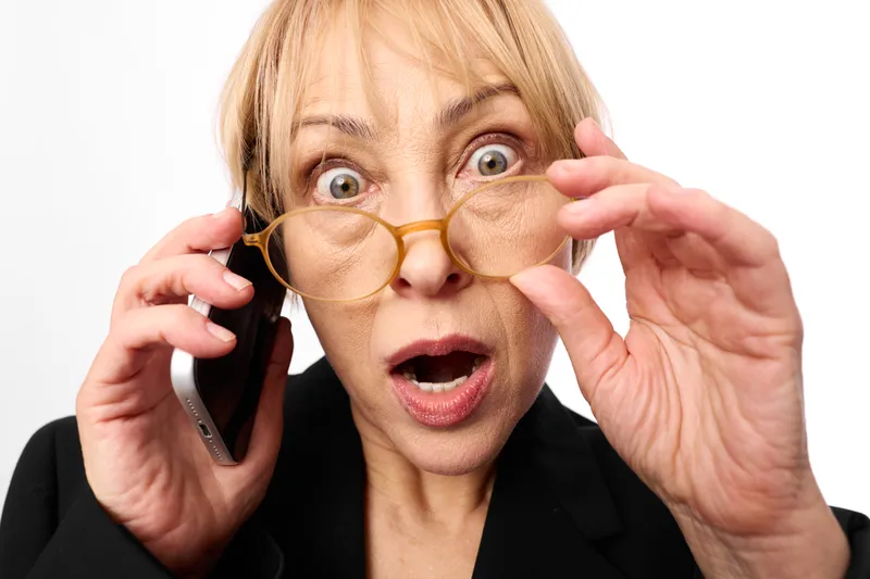 Surprised senior woman holding a phone to her ear, wearing glasses, eyes wide and mouth open, adjusting frames with one hand, showing shock and stress, close up on background