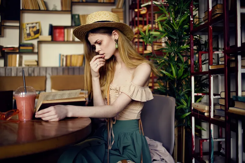 Женщина в шляпе сидит в кафе, у нее на столе напиток и книга, она читает и отдыхает в отпуске