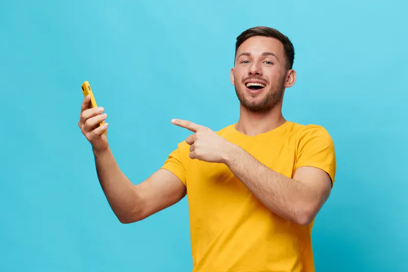 Привлекательный загорелый мужчина в жёлтой футболке с улыбкой указывает пальцем на смартфон, позируя изолированно на синем студийном фоне