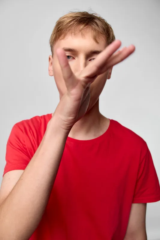 Hands covering part of face on a close up of a boy in a red shirt, conveying concealment, privacy, and selective reveal for dramatic, expressive storytelling in studio lighting