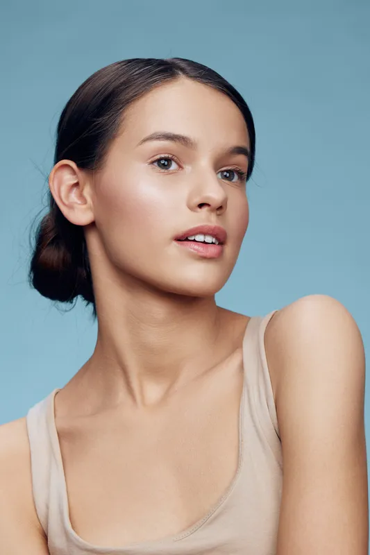 Young woman with natural makeup and a neutral tank top, showcasing confidence and radiance against a light blue background