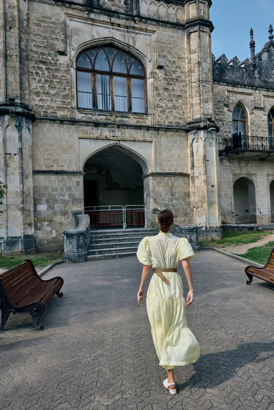 Woman in a pale yellow dress walks toward a historic castle entrance in a sunlit courtyard with ivy clad walls and ancient arches overhead, capturing a timeless travel moment.