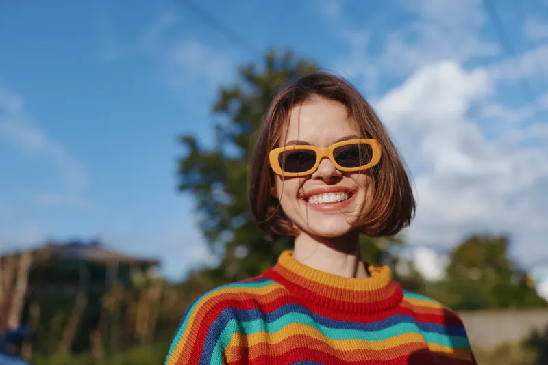 woman smile sunglasses sweater portrait outdoors happy colorful young adult in rainbow knit pullover, short hair and bright accessory under blue sky, lifestyle and joyful expression