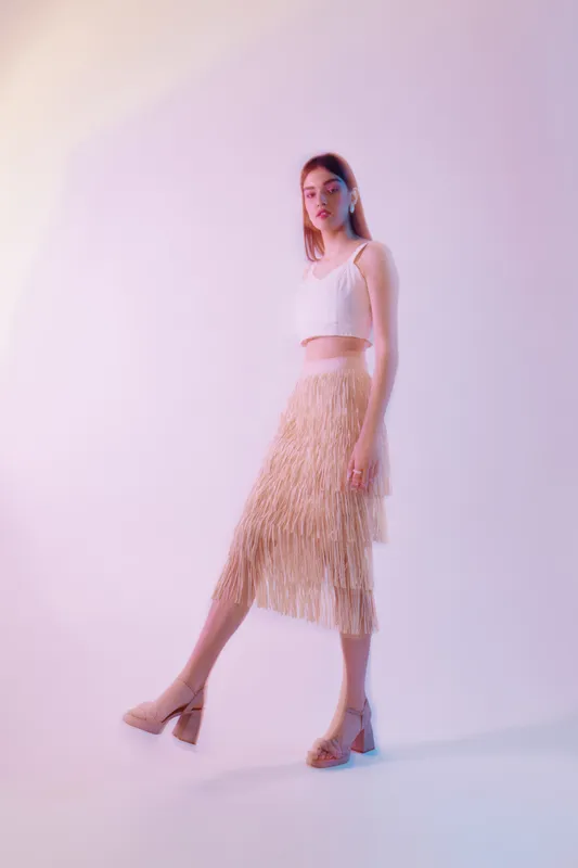 Young woman in a white crop top and beige fringed skirt poses confidently in a studio with soft pastel lighting. Concept of woman beauty fashion and modern style.