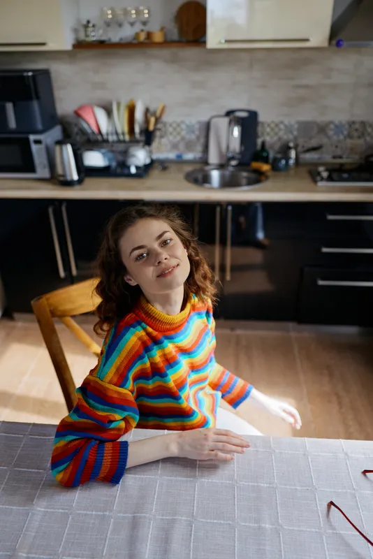Casual woman sits at a kitchen table wearing a colorful striped sweater, enjoying a relaxed morning at home, candid lifestyle moment, warm natural light, cozy domestic scene.