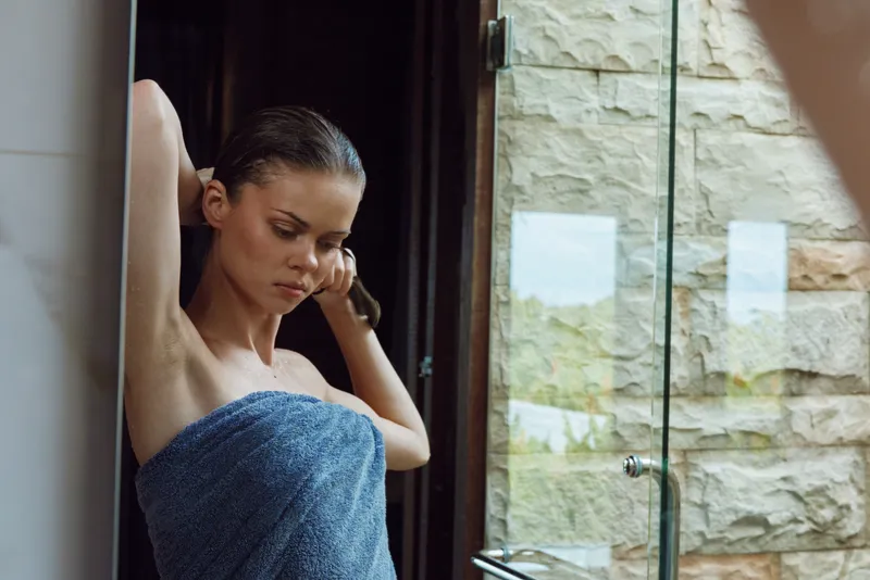 young woman wrapped in blue towel adjusting hair indoors near glass window with stone wall background, natural light highlighting calm and thoughtful expression