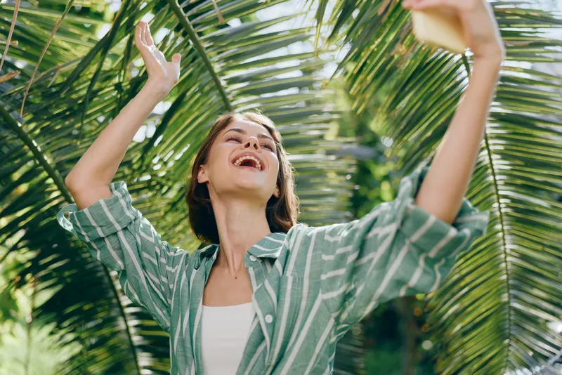 A joyful woman raises her arms outdoors among palm leaves, smiling broadly in bright daylight.