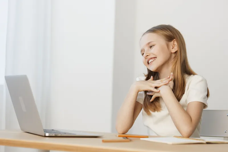 Young smiling girl interacting with a laptop, showcasing joy and enthusiasm, in a bright, minimalistic home office setting, emphasizing modern technology and learning