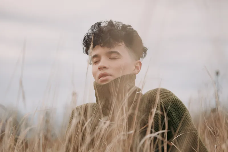 Portrait of a thoughtful youth in a windlit field, wearing a chunky knit turtleneck, eyes closed, conveying authenticity and credibility through natural pose and honest mood.
