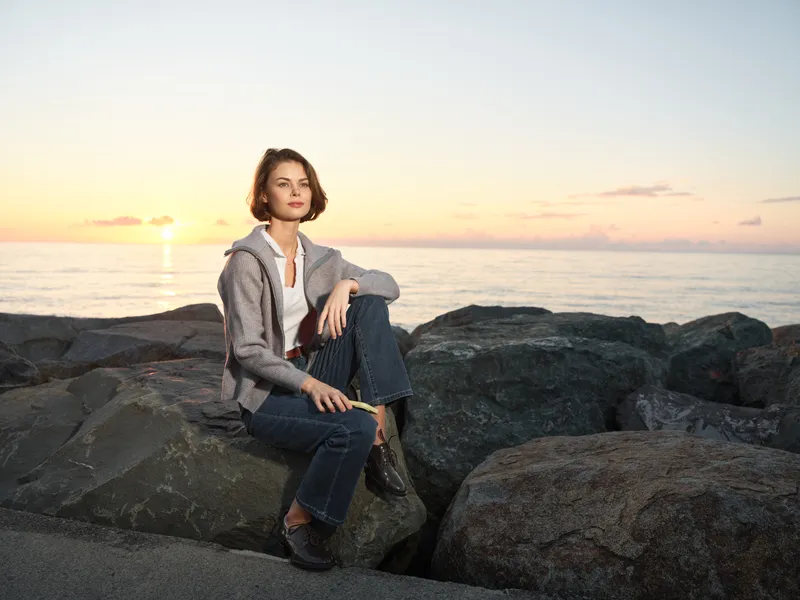 A calm woman sits on rocky shoreline during sunset, wearing a casual jacket and jeans. She gazes toward the water with a thoughtful expression, creating a peaceful and reflective mood along the coast.