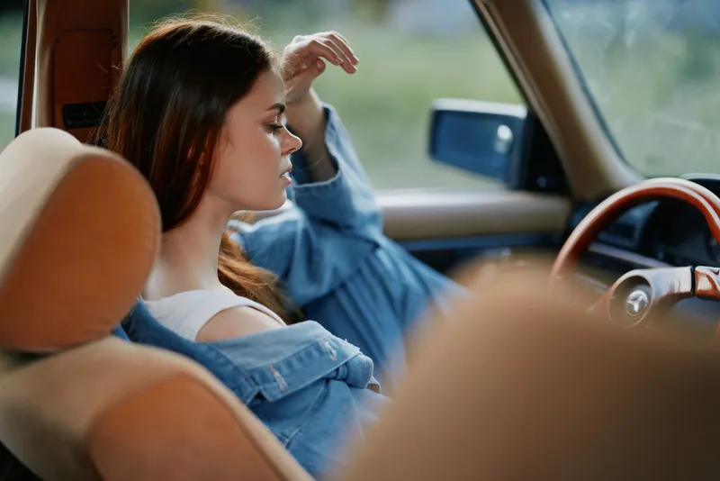 Thoughtful young woman in denim jacket sitting in vintage car, showcasing emotions of reflection and nostalgia with natural background.