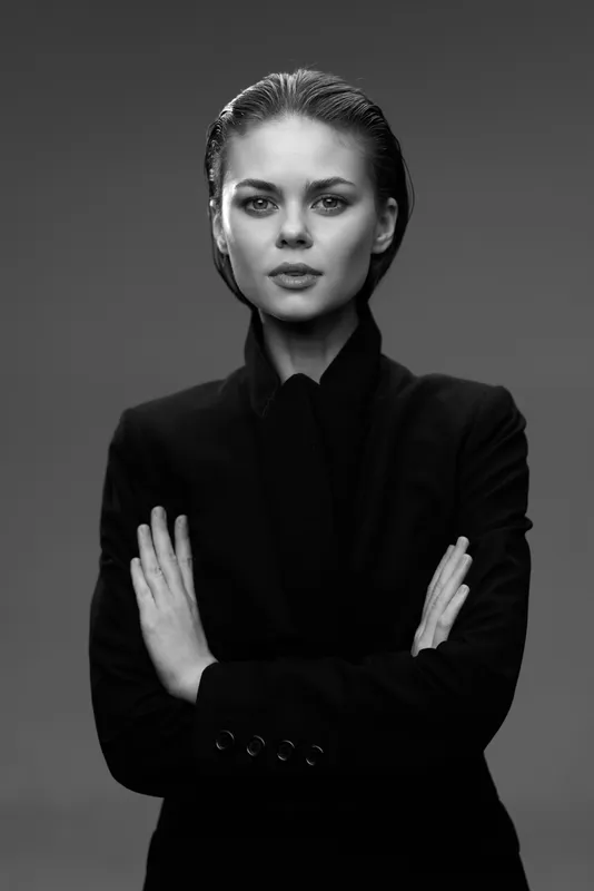 Black and white portrait of serious woman in stylish black clothing with crossed arms and intense gaze.