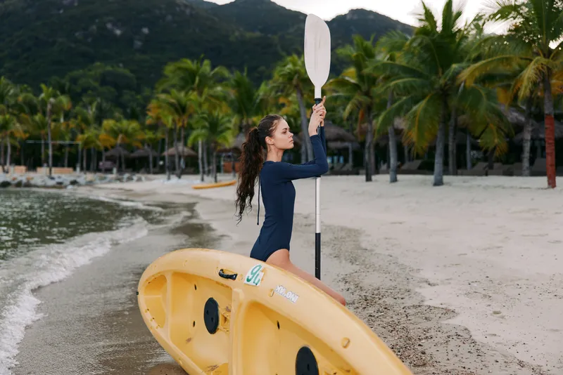 Young woman on a sandy beach lifts a paddle beside a bright yellow kayak, framed by palm trees and calm water, capturing seaside adventure and carefree outdoor activity.