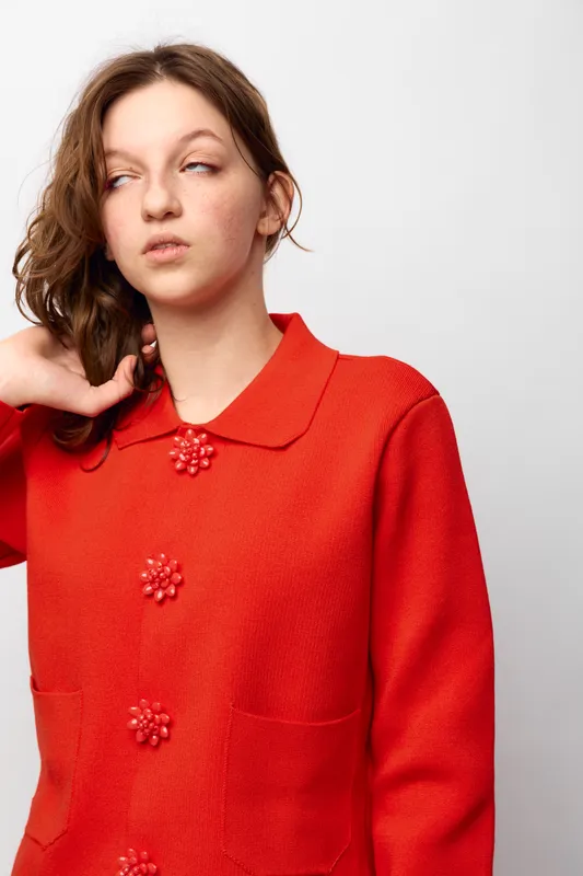 Young woman in red sweater with decorative buttons looks sideways with thoughtful expression. Studio portrait on white background. Concept of fashion, casual style, and contemplation.