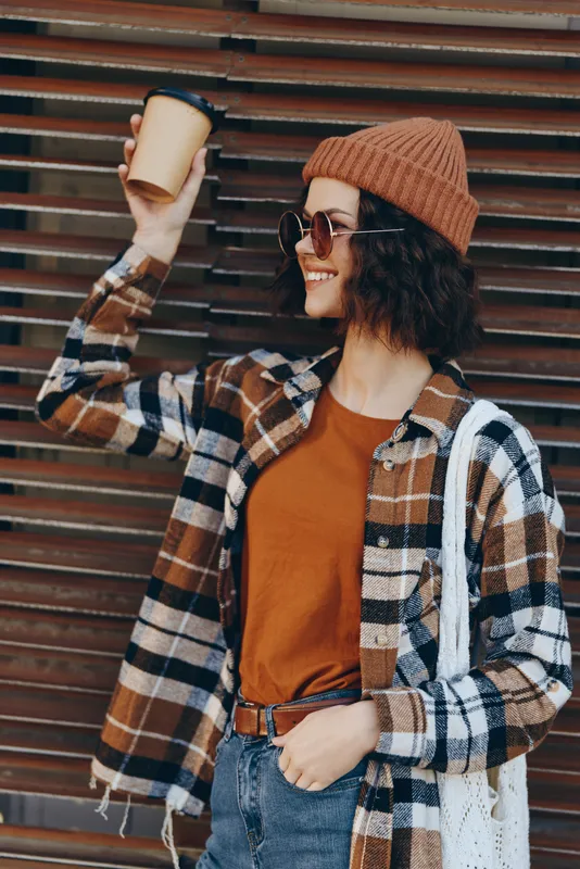 woman coffee smile urban beanie plaid sunglasses autumn. Candid lifestyle portrait of a smiling woman holding coffee cup on city street with golden hour glow, authenticity and emotional storytelling.