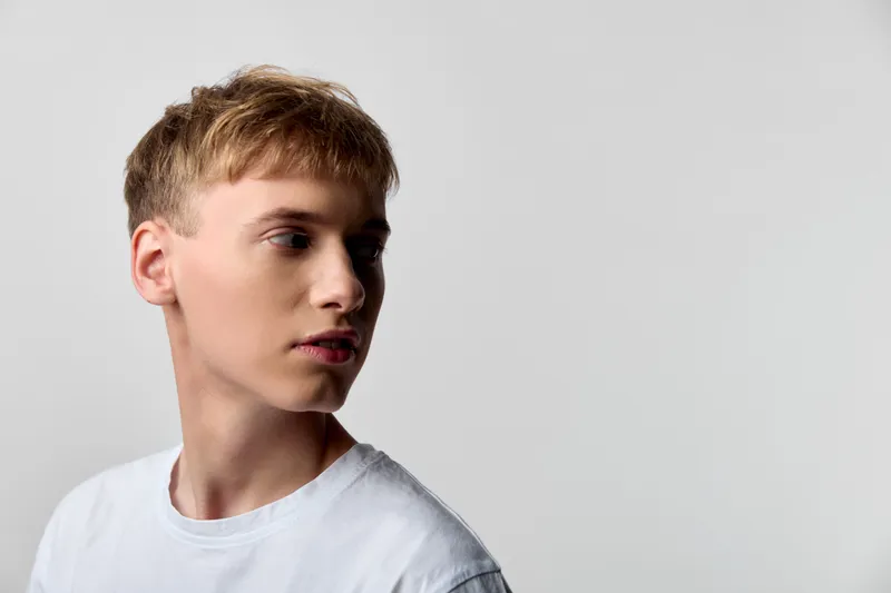 Young man with a calm face in profile, neutral expression, lit by studio light, wearing a white tshirt for a clean headshot style against a minimal background.