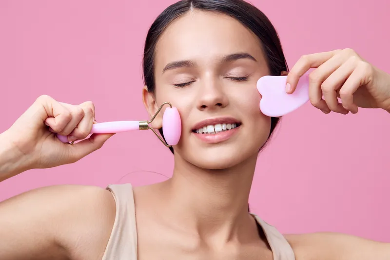 Young woman enjoying skincare with a facial roller on a pink background, promoting healthy skin and self care routines