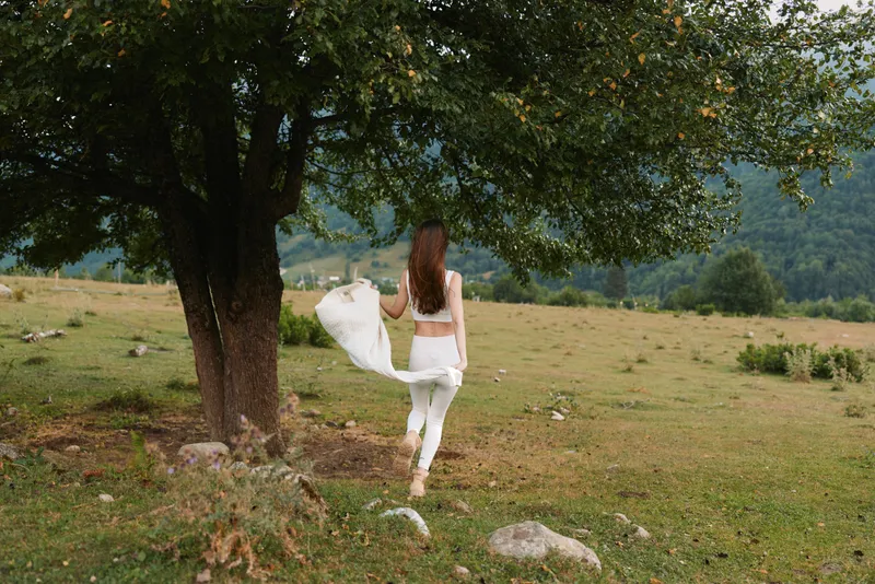 Young woman with long brown hair walks barefoot on grass near a large tree in a peaceful meadow surrounded by hills and greenery on a cloudy day.