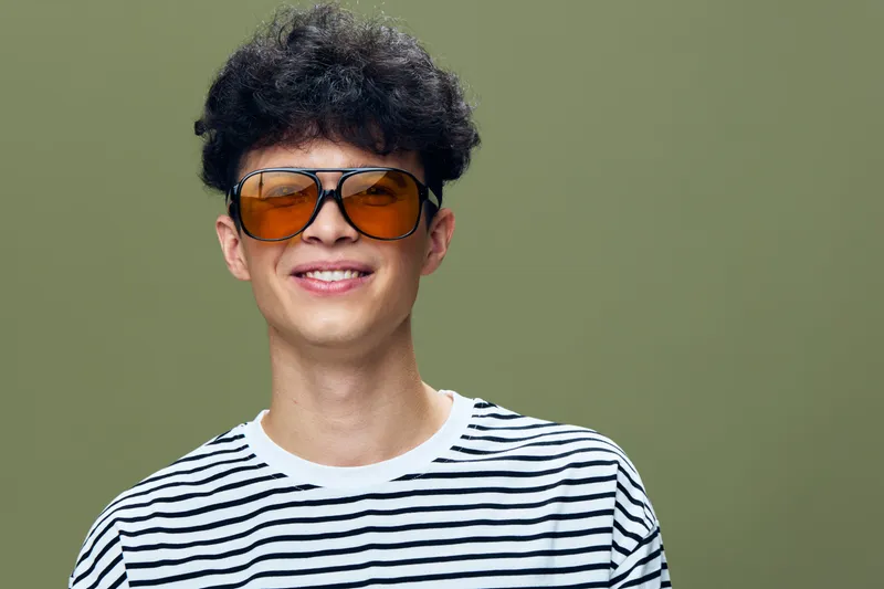 smiling young man with curly hair wearing large orange sunglasses and striped shirt, green background, casual style, positive expression, studio portrait