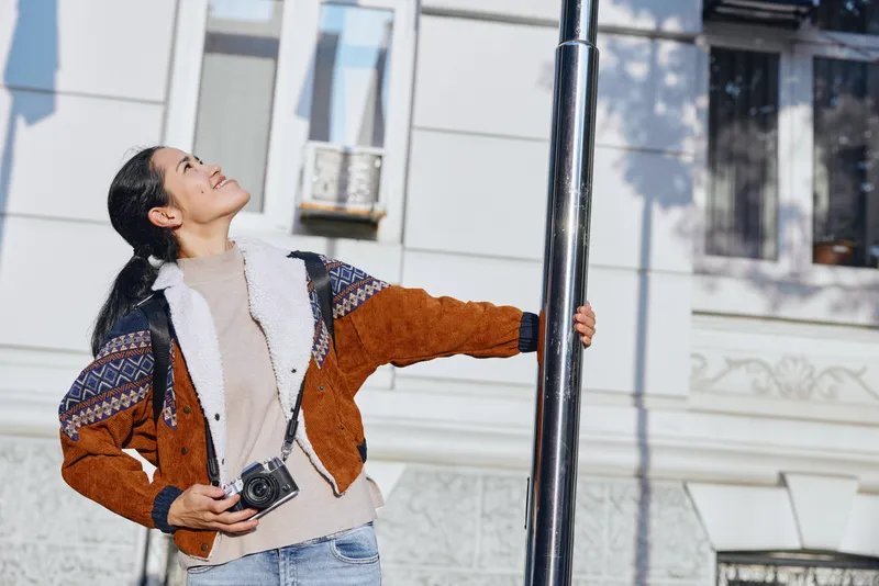 Woman wearing a chunky knit jacket and scarf, holding a camera, gazing up at a street lamp on a sunny urban street, capturing casual travel style and a candid moment