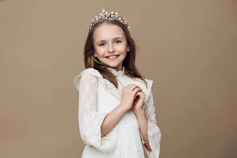Charming young girl in a white dress with a crown, smiling joyfully against a neutral beige background, representing childhood innocence and beauty