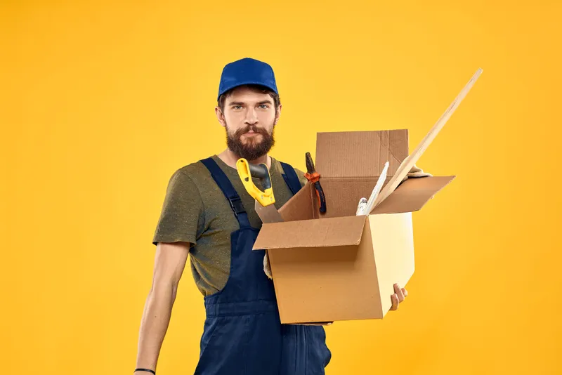 Мужчина в рабочей форме с коробкой, загруженной инструментами, на жёлтом фоне